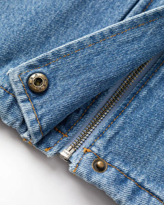 Détail en gros plan d’un jean en denim bleu stone fade, 100 % coton, avec une fermeture éclair métallique et des boutons rivets apparents, surpiqûres orange contrastantes.