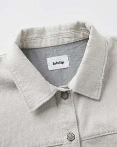 Col chemise en denim non teint couleur granit clair avec coutures apparentes et boutons métalliques, tissu 100% coton au toucher structuré et naturel.