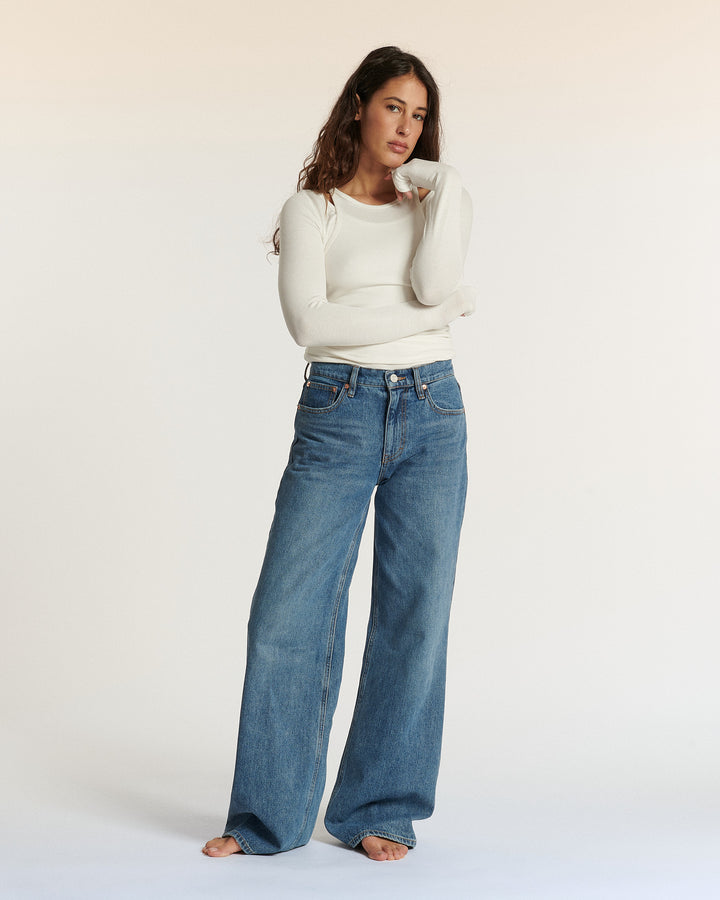 Femme portant un jean large en denim bleu délavé, associé à un haut blanc à manches longues, posant dans un studio à fond clair.