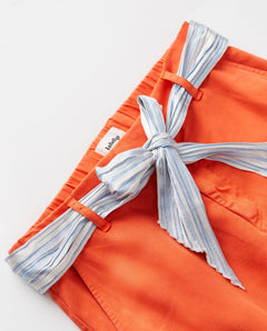 Jean femme en tissu Tencel orange vif, coupe droite avec ceinture nouée en tissu rayé bleu et blanc, style estival et confortable adapté à toutes morphologies.